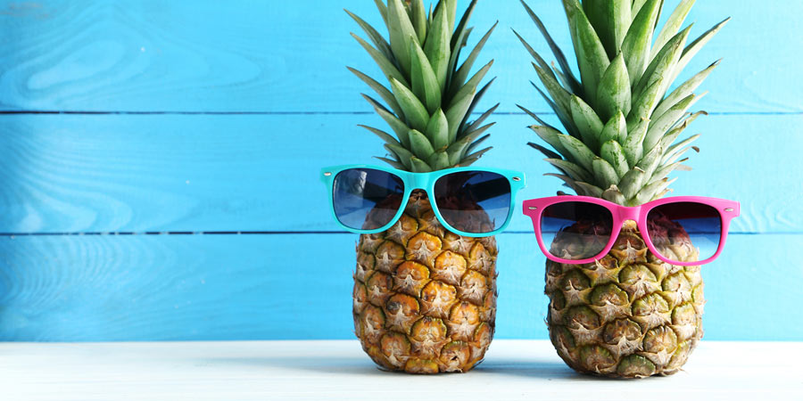 Two pineapples against a bright aqua background wearing plastic sunglasses in blue & pink to imply they are swingers